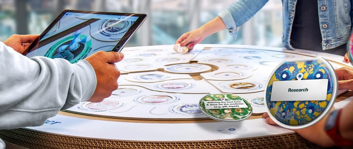 A player holds a tablet over the Make Your Mark board, its screen showing the board in augmented reality, while others place discs such as 'Research'