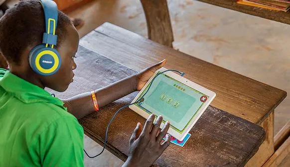 A child in Uganda wearing headphones plays a Can't Wait To Learn maths exercise on a tablet at a wooden school desk